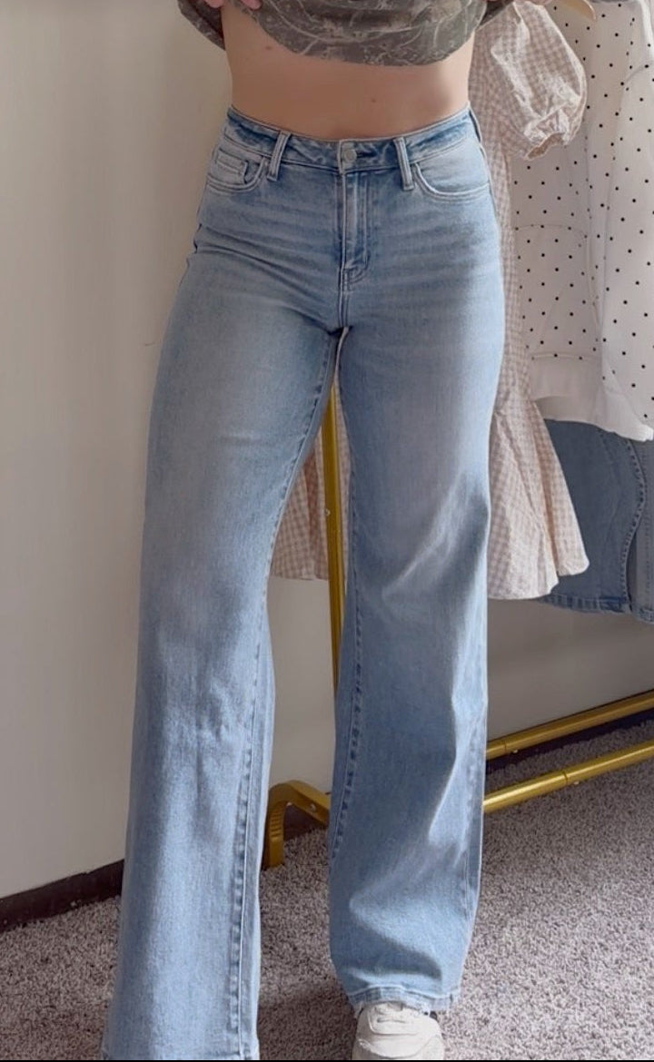 A woman stands in a room wearing a green camouflage cropped sweatshirt with 'PRAY' printed on the front and light blue wide-leg jeans. A clothing rack with other garments is visible behind her next to a framed floral painting.